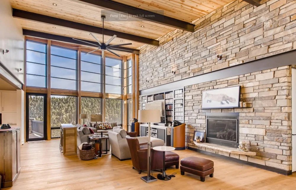 Great room with a stone fireplace wall, timber ceiling and tall windows looking out to the trees